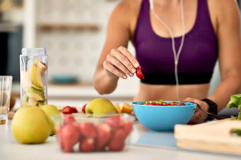 mulher prepara um pré treino em uma mesa de vidro com algumas frutas, como morango e maçã verde.